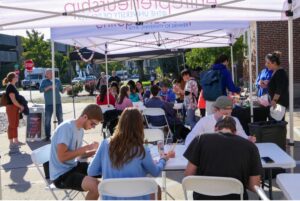 Open house guests sit under a tent at a table crafting