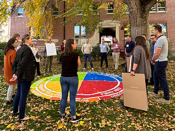 ALX participants stand around a giant, four-colored Insights Discovery Wheel outside of a DU building